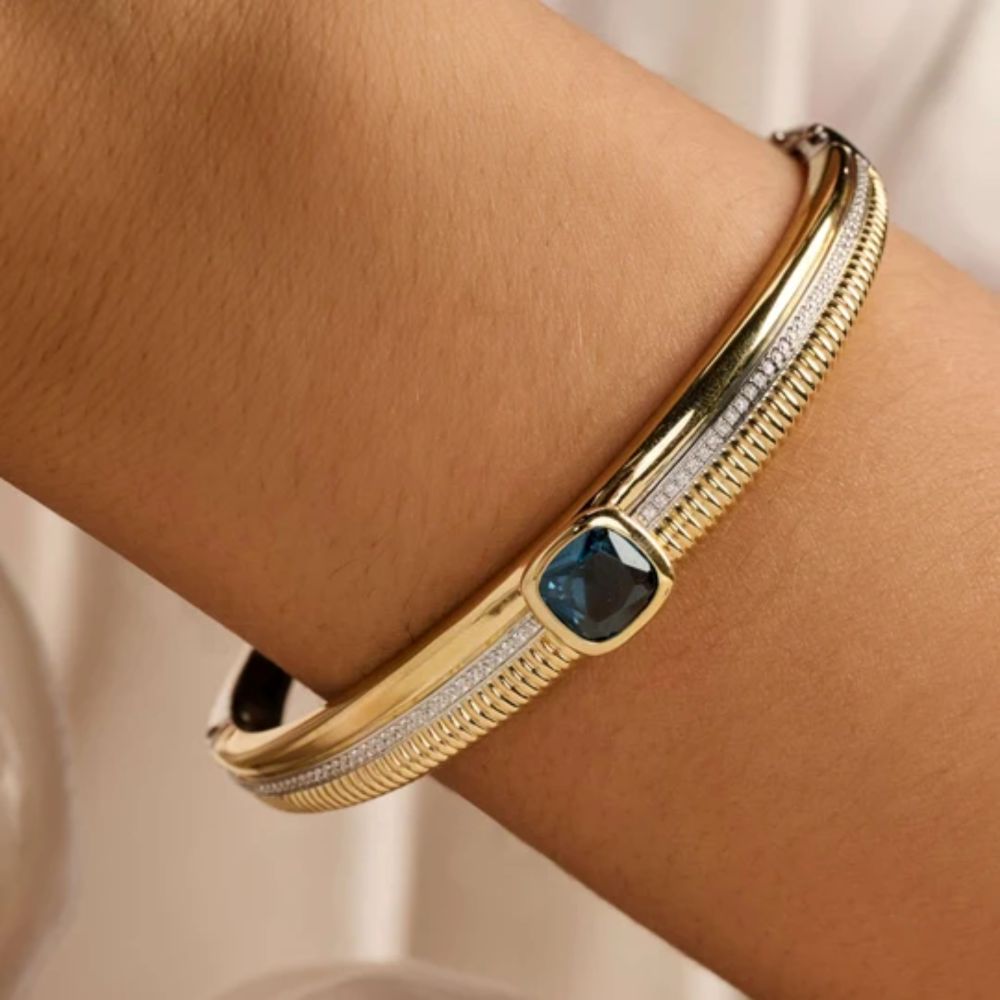 A close-up of a gold bangle bracelet with a textured channel, a row of small clear stones, and a central square dark blue gemstone, shown worn on a person's wrist.