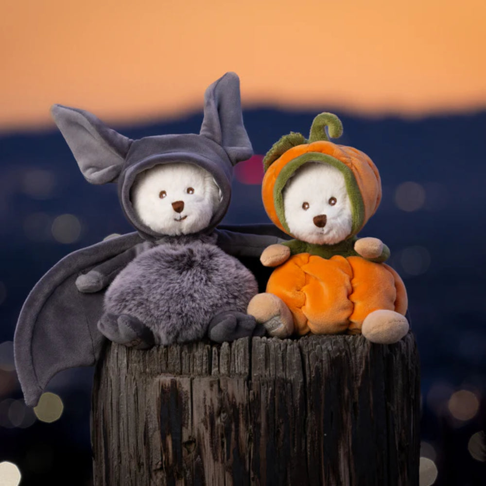 Two white teddy bears, one in a bat costume and the other in a pumpkin costume, sitting on a wooden stump against a blurred background.
