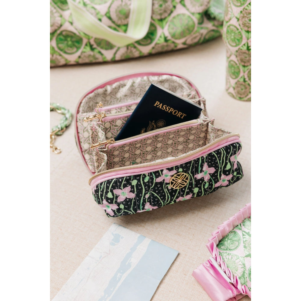 A small zippered pouch with a black floral exterior, pink trim, and a beige monogram interior, shown open with a passport inside. The pouch is photographed on a light beige surface with other matching patterned accessories partially visible around it.