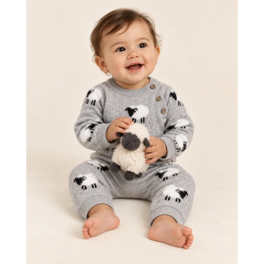 A smiling baby sitting on a cream studio background wearing a gray knit one-piece jumpsuit with black and white sheep prints and holding a small plush sheep toy.