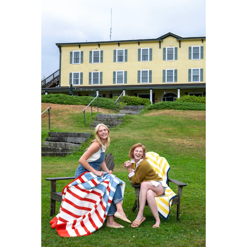 Two women sitting on wooden chairs with colorful blankets in front of a yellow building.