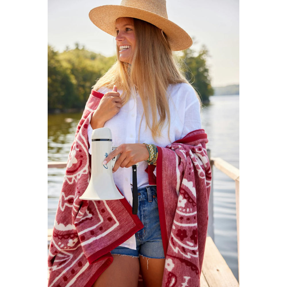 Woman on a dock by a lake, wearing a straw hat and red patterned blanket, holding a white sound device. 