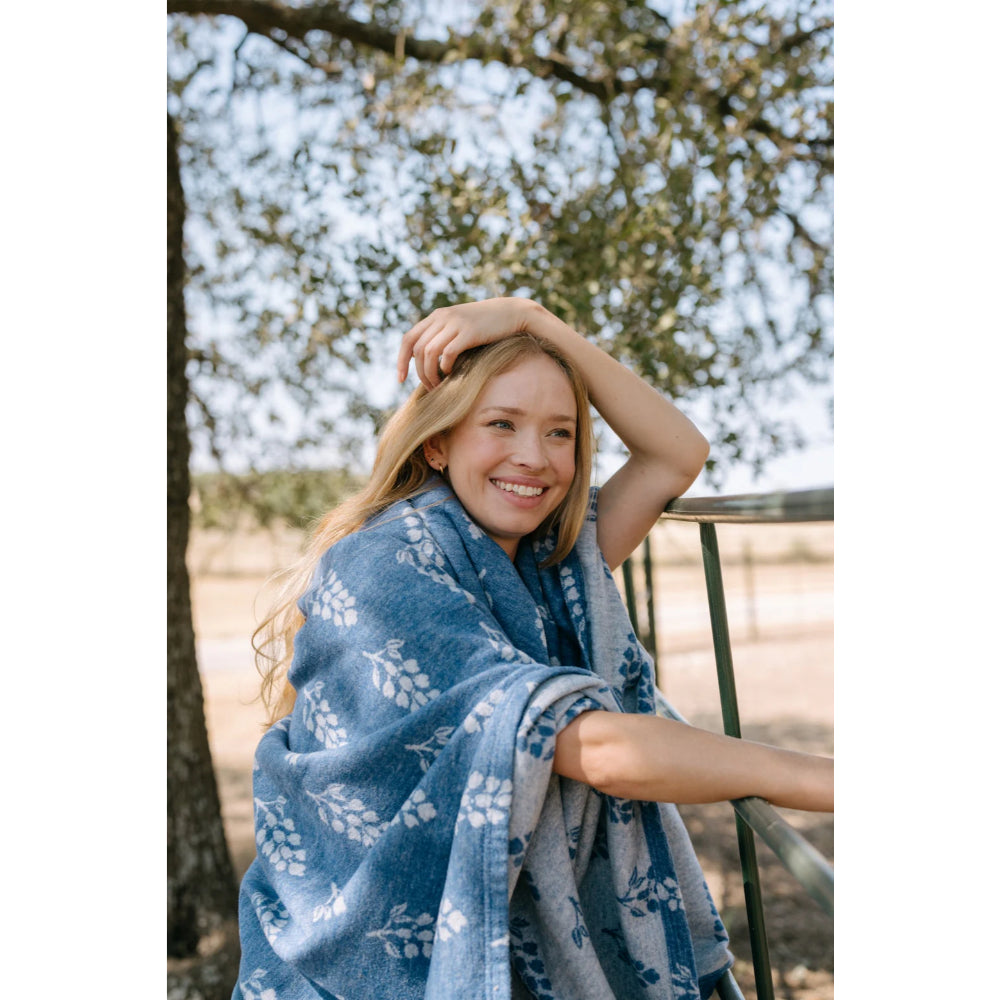 A woman wearing a lightweight blanket around her with blue colors and white floral patterns in an outside background. 
