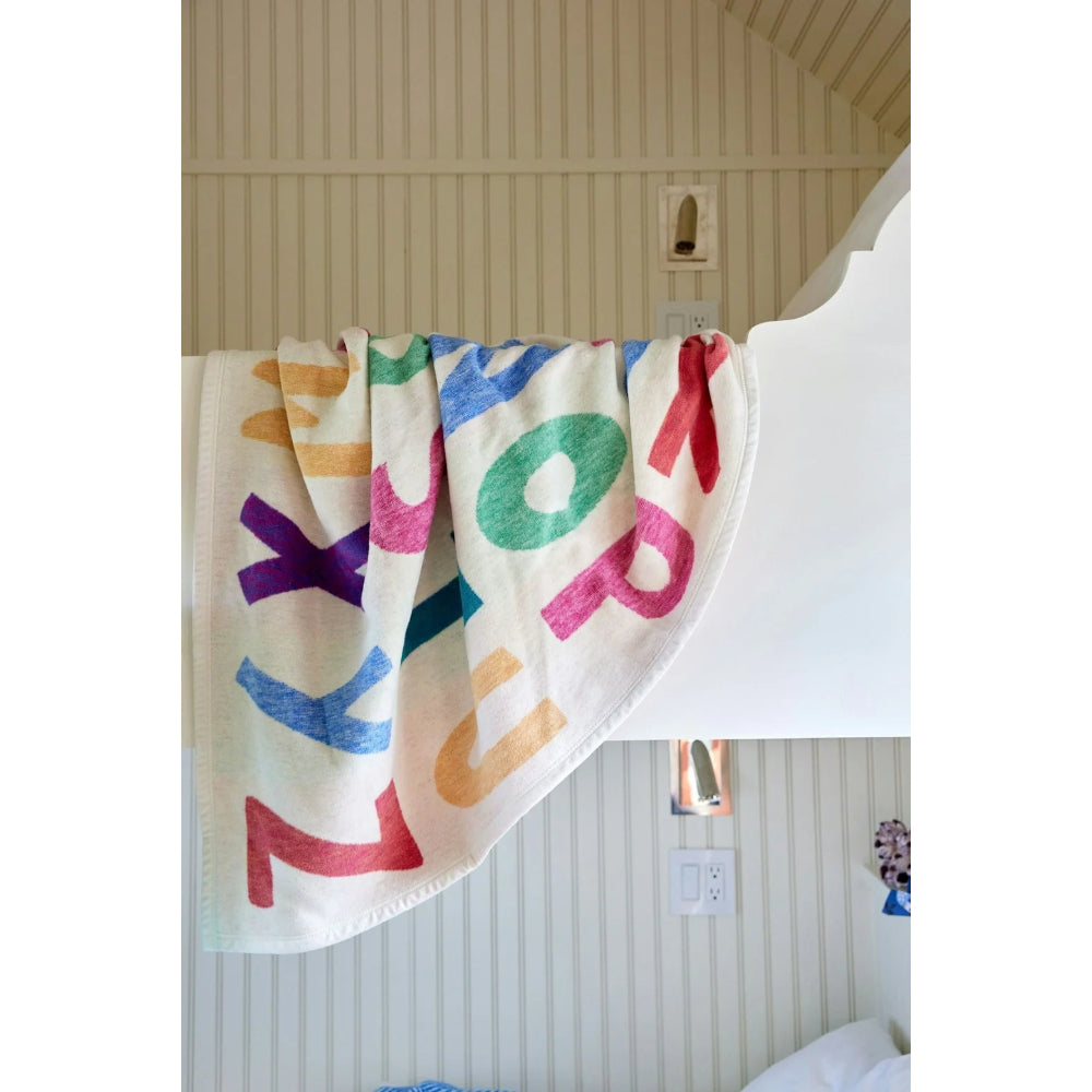 A cream-colored cotton blanket featuring large, colorful hand-drawn style letters spelling 'ZOO' in pink, blue, green, orange, and purple, draped over the side of a white bed in a bedroom with beige paneled walls.
