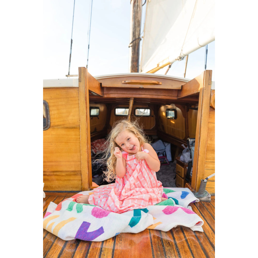 A young girl with curly blonde hair sits smiling on the wooden deck of a sailboat, wrapped in a pink and white patterned blanket or dress. She is sitting on a white blanket with colorful pastel shapes.