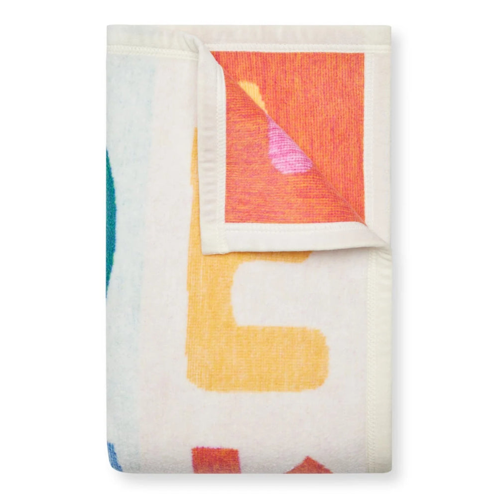 A folded throw blanket with a cream base and a colorful abstract geometric pattern, featuring a folded corner that reveals a textured orange and pink inner side.