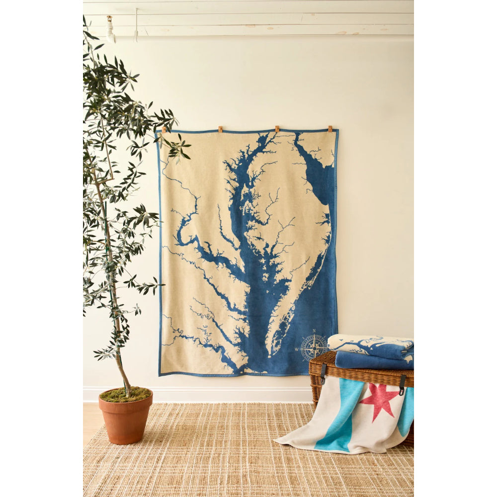A large woven tapestry featuring a blue waterway or tree-like pattern on a cream background, hanging on a wall in a styled room with a potted plant, woven rug, and a bench with folded textiles.