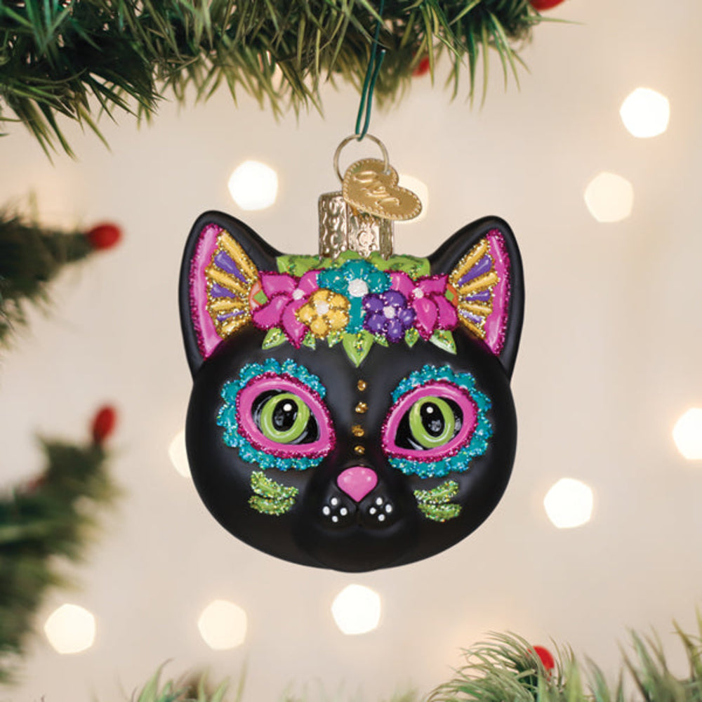 A black cat-shaped Christmas ornament decorated with colorful floral patterns and glittery eyes, hanging from a green tree branch with warm bokeh lights in the background.