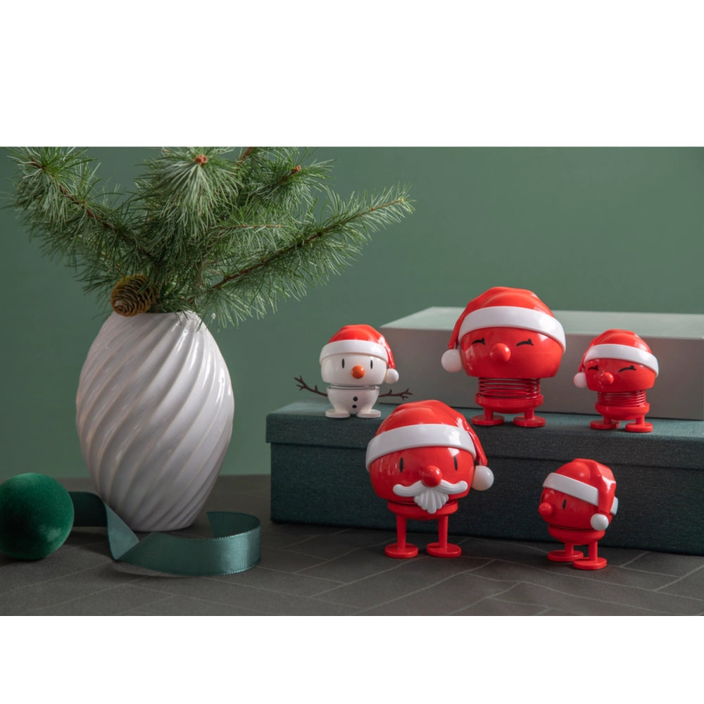 Five red cartoon Santa Claus-style figurines in different poses, displayed on a dark gray box next to a white vase with pine branches and a green ribbon against a muted green wall.