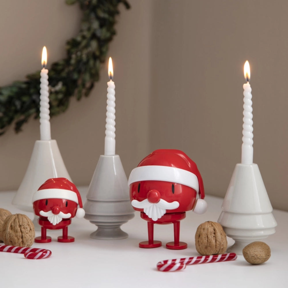 Three Santa Claus-shaped candle holders with red bodies, white hats and beards, each holding a white spiral candle. The scene is styled on a tabletop with walnuts, candy canes, and a green wreath in the background.