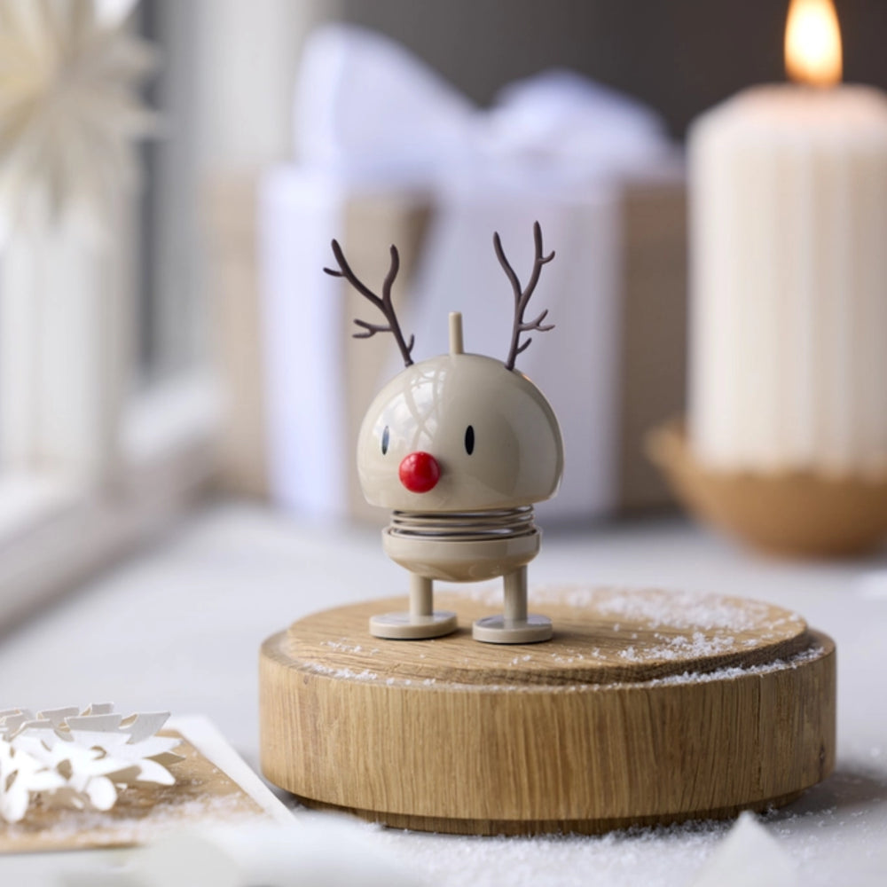 A small holiday reindeer figurine with antlers and a red nose stands on a round wooden base with a dusting of faux snow, set in a softly blurred festive interior with a candle and wrapped gift in the background.