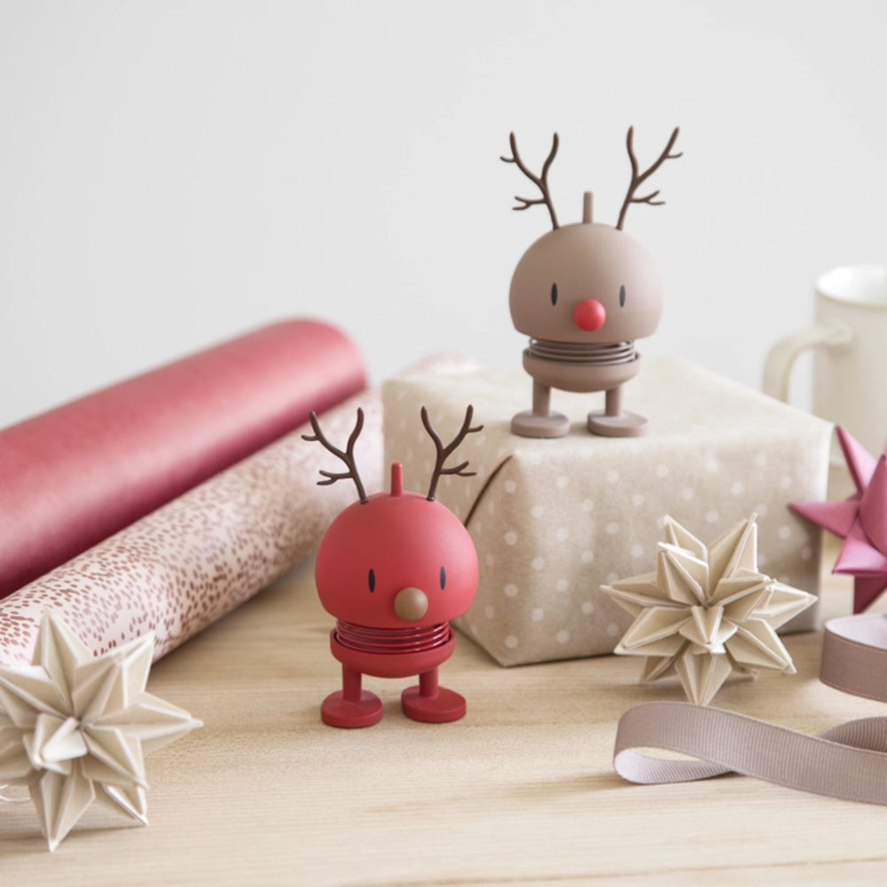 Two small reindeer figurines with antlers and spring-loaded legs, styled on a tabletop with wrapping paper, origami stars, and a mug in the background.