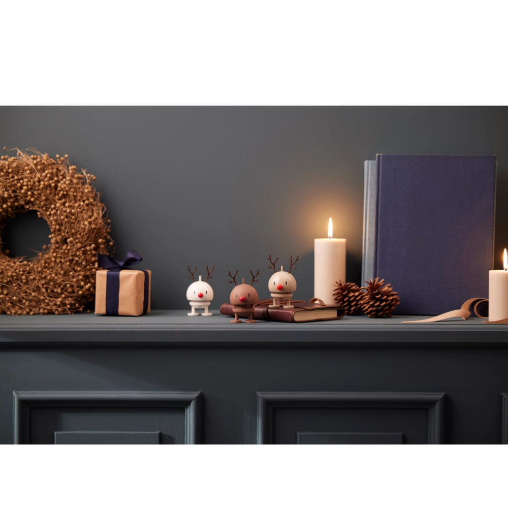A styled holiday mantel scene with a dark gray wall and matching cabinet. Decorative items include a dried floral wreath, a wrapped gift box, two small reindeer figurines, candles, pinecones, and a navy book or box.