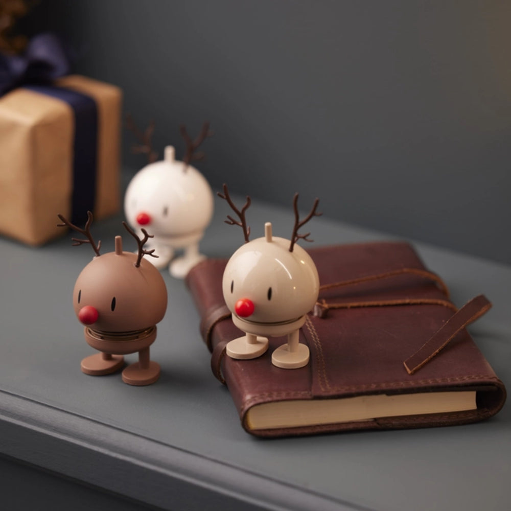 Three small reindeer-shaped decorative figures with antlers and red noses are arranged on a gray tabletop beside a brown leather notebook, with a wrapped gift box blurred in the background.