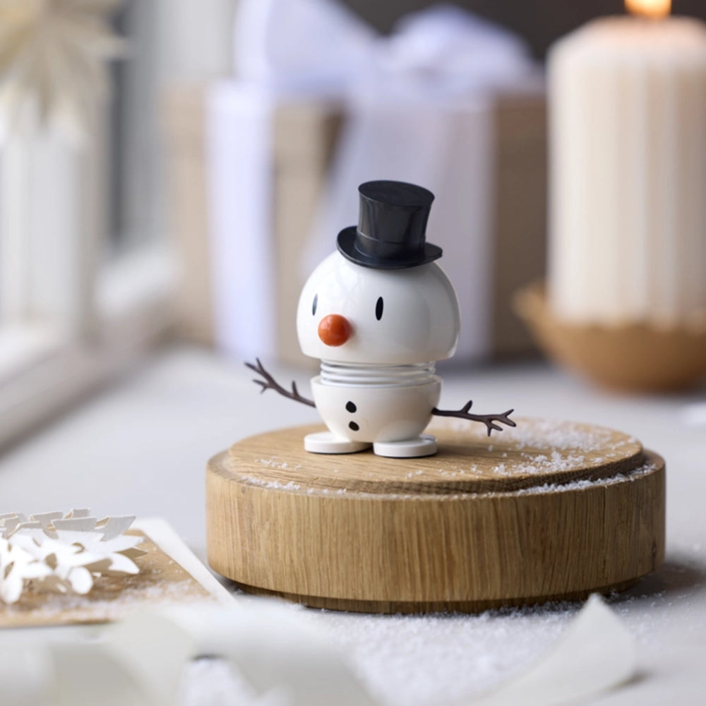 A small white snowman figurine wearing a black top hat and orange carrot nose, standing on a round wooden base with a light dusting of faux snow, photographed in a softly blurred holiday setting with candles and wrapped gifts in the background.