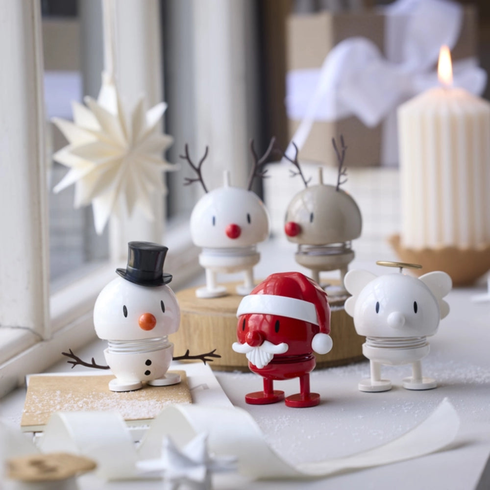 A group of small white holiday figurines arranged on a tabletop near a window: a snowman with a black top hat, a Santa-style red figure with white beard, two reindeer figures with antlers, and a white mouse-like figure with a halo. A lit candle and soft home decor are blurred in the background.