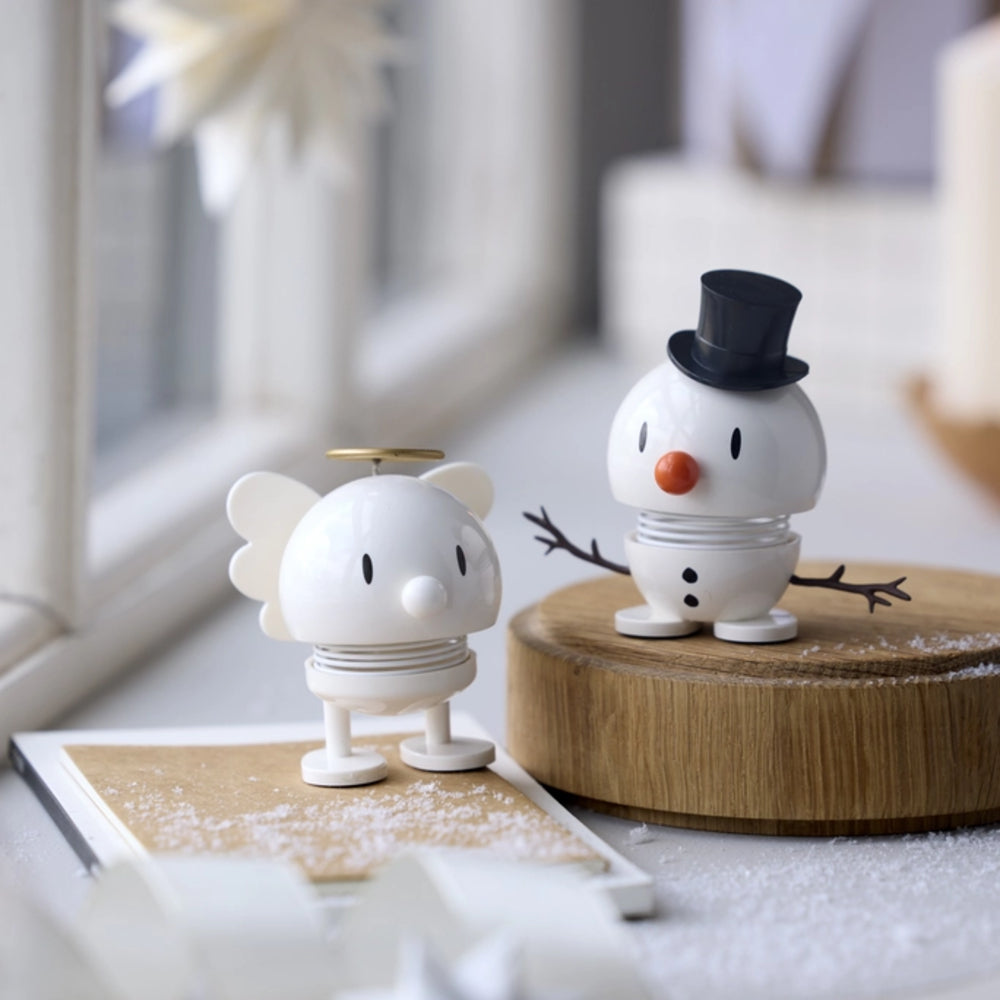Two small decorative figurines styled like a white mouse with a gold halo and a white snowman wearing a black top hat, displayed on a tabletop with a blurred window and soft indoor background.