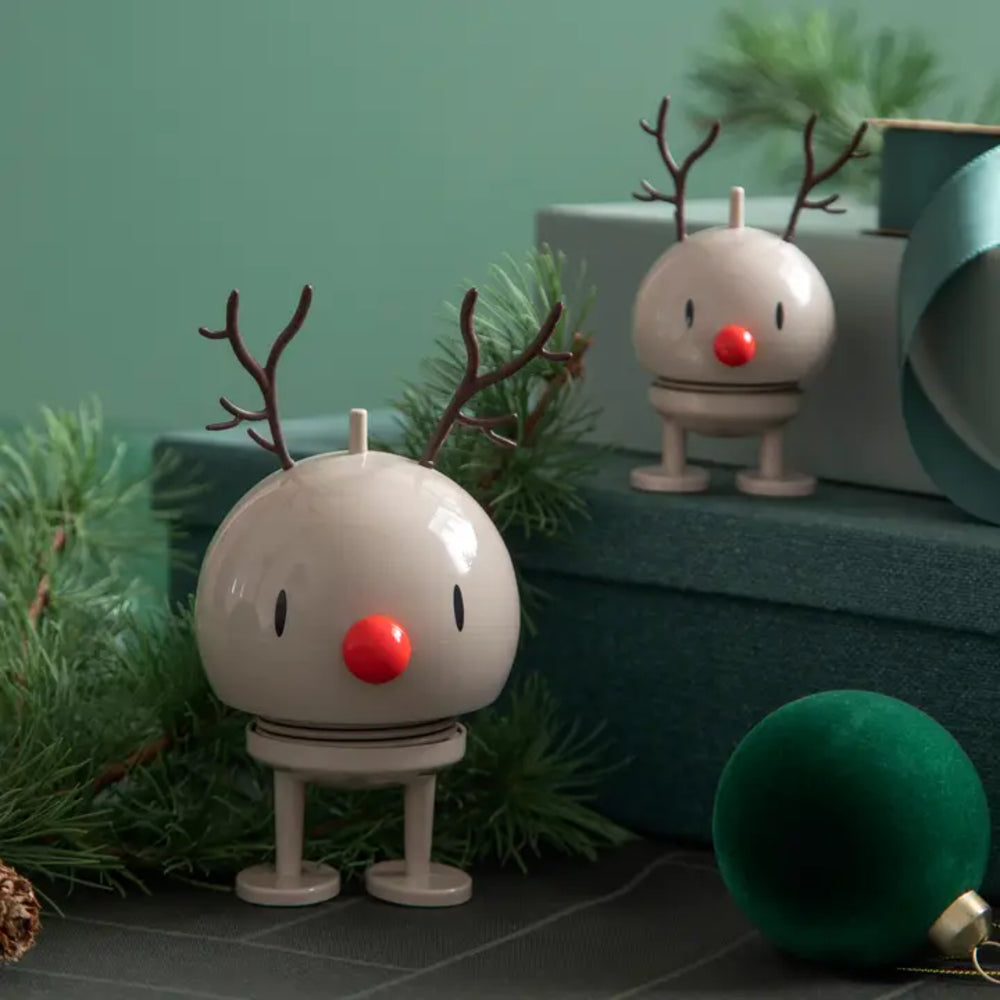A festive holiday decoration shaped like a reindeer, featuring a glossy off-white round head, brown antlers, a bright red nose, and small legs. It is styled on a dark surface with pine branches, a green ornament, and a second matching reindeer decoration in the background.