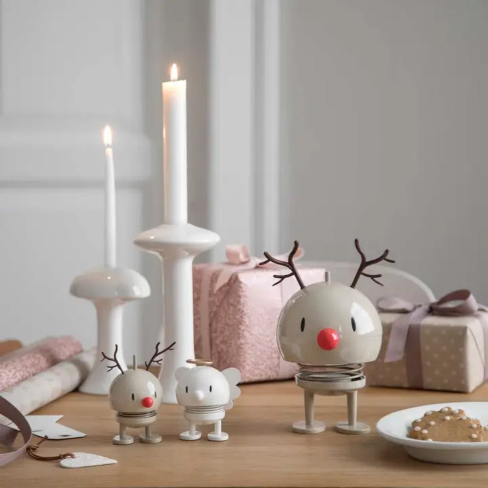 Three small white reindeer-shaped spring toys with antlers and red noses are arranged on a wooden table beside two lit white candles, pink gift boxes, and a plate of candy.
