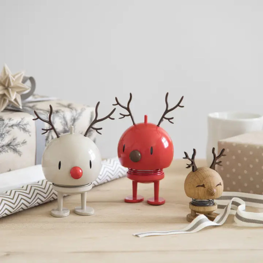 Three small reindeer figurines styled on a light wooden tabletop with gift boxes and ribbons in the background. The left reindeer is cream with a red nose, the center reindeer is bright red, and the right reindeer is natural wood.