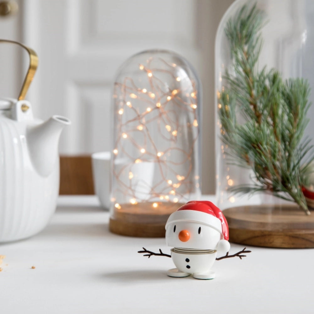 A small white snowman figurine wearing a red Santa hat stands on a white tabletop in a softly styled holiday setting with glass domes, warm string lights, pine branches, and a white teapot in the background.