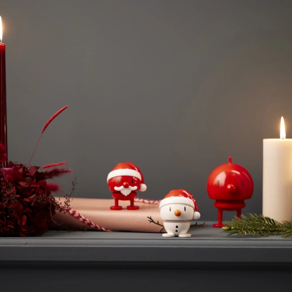 A holiday-themed tabletop scene with two small decorative figures: a Santa Claus figure and a snowman figure, both wearing red Santa hats. There is also a glossy red ornament-shaped object on a stand.