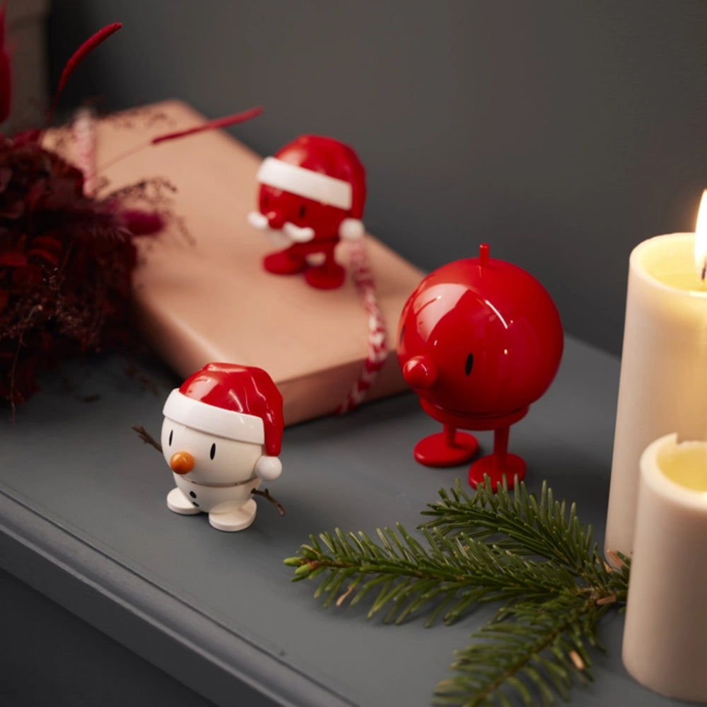 Three small red-and-white Christmas figurines displayed on a gray tabletop with a wrapped gift, evergreen sprigs, and lit white candles in a cozy indoor setting.