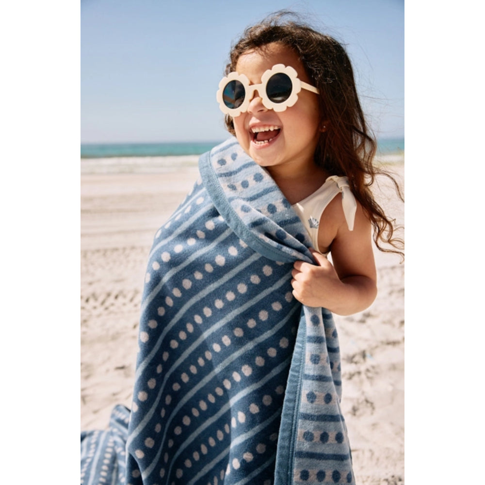 A young child wrapped in a blue patterned beach towel on a sandy beach, wearing white flower-shaped sunglasses.