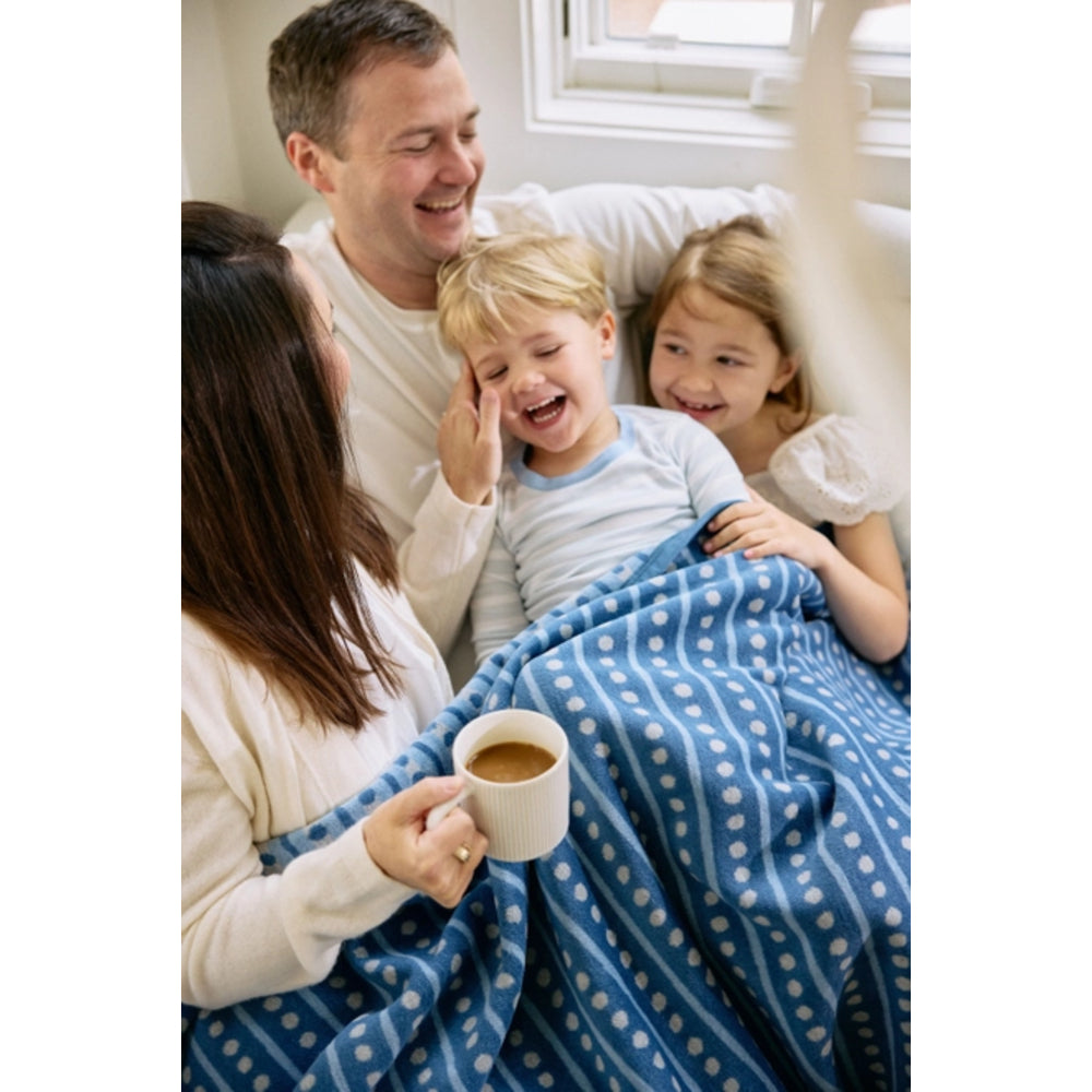 A family of four sharing a cozy moment under a blue patterned blanket in bed. The mother holds a white mug, while the father and two children are smiling and laughing.