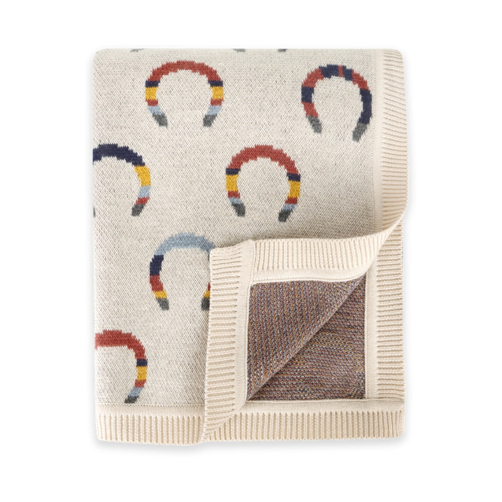A cream-colored knit blanket featuring a repeating pattern of colorful horseshoes, with ribbed borders and a folded corner showing a textured brown lining.