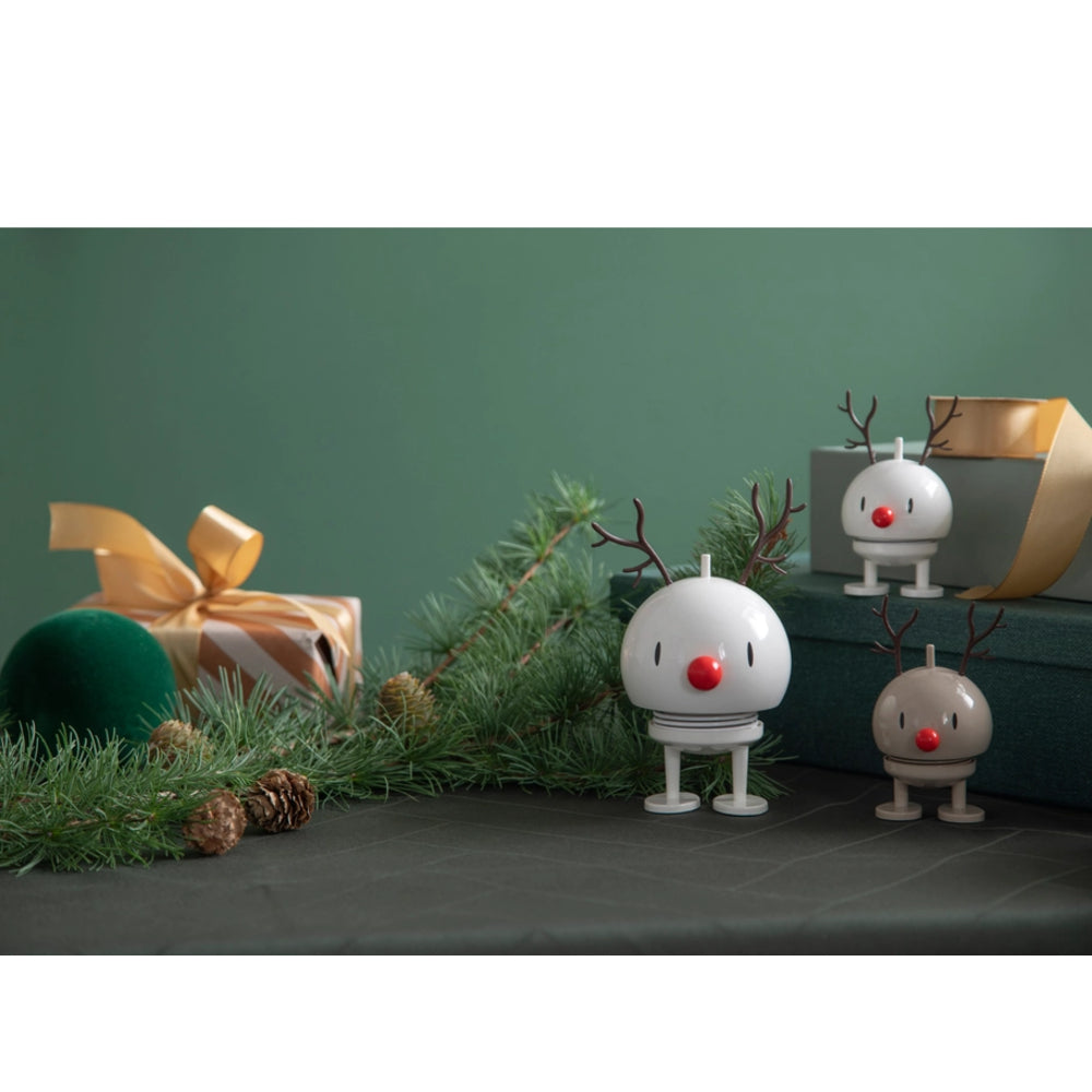 Three white reindeer figurines with red noses and brown antlers, styled with pine branches, pinecones, and wrapped gifts against a dark green background.