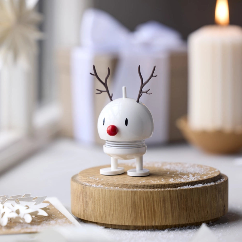A small white holiday reindeer figurine with brown antlers and a red nose stands on a round wooden base with a light dusting of snow. The scene is softly lit with a blurred gift box and a lit candle in the background.