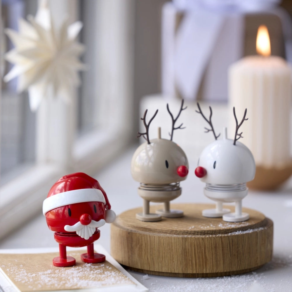 A festive tabletop display with a small red Santa figure on the left and two small reindeer figures on a round wooden base to the right, styled near a lit candle and soft window light.