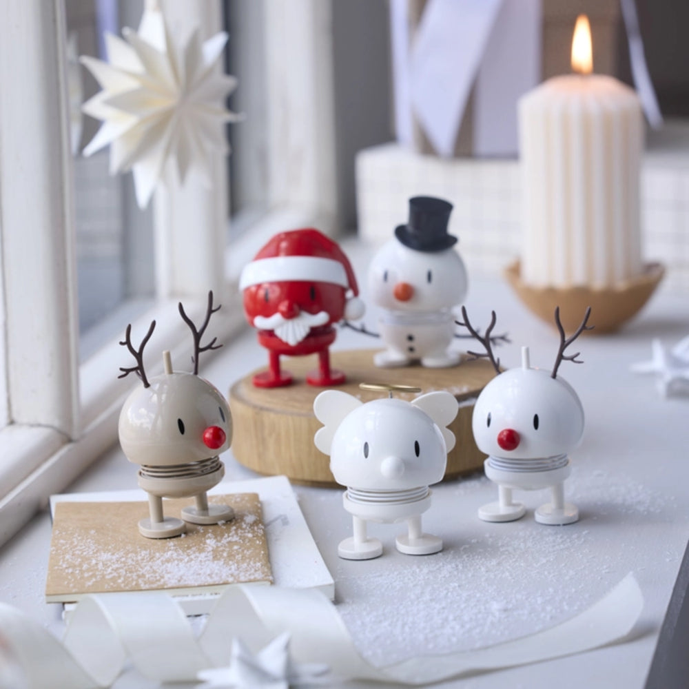 Three small holiday figurines displayed on a tabletop near a window: two reindeer with antlers and red noses, a white snowman with a black top hat, and a Santa Claus figure in red. A lit candle and white decorative paper stars are in the background.