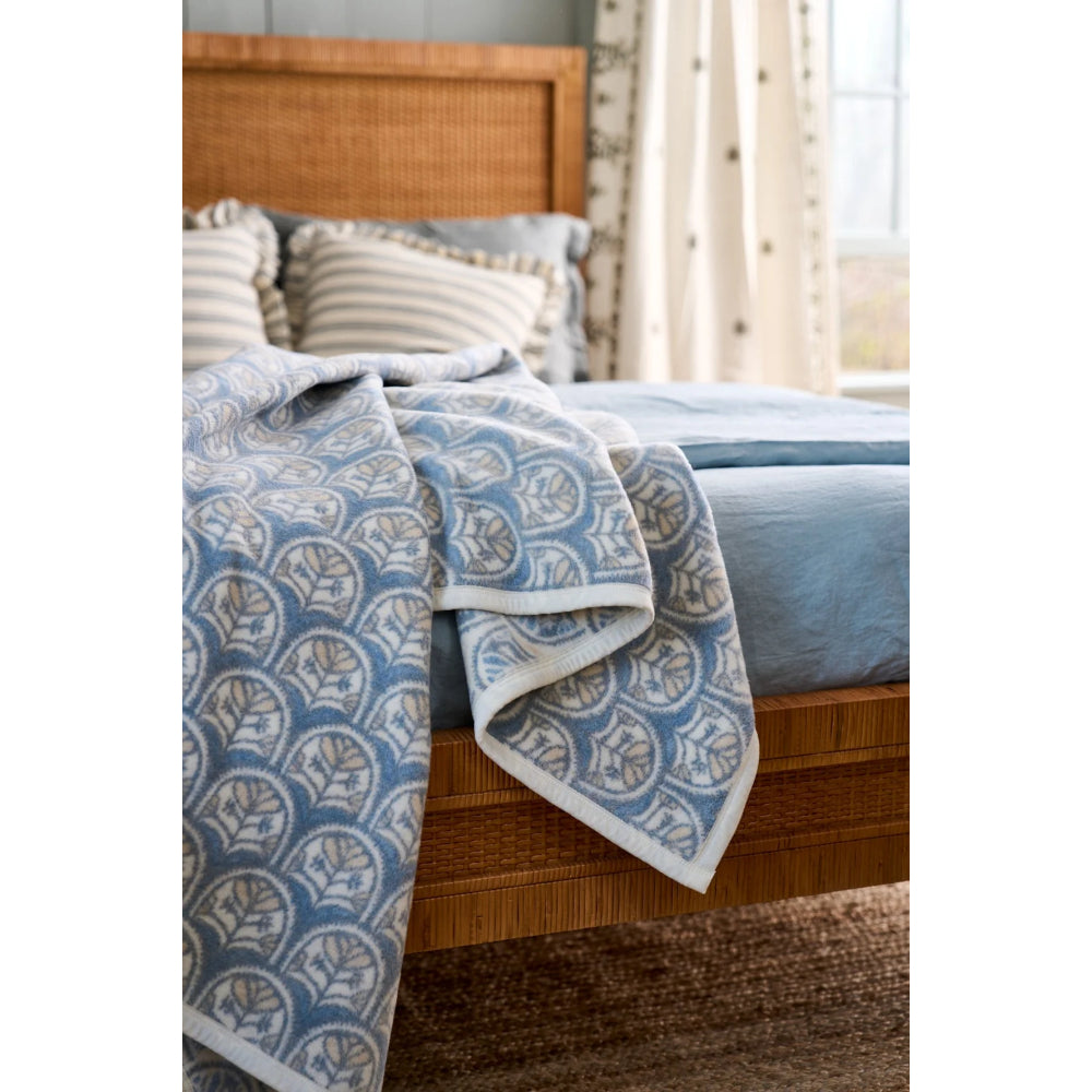 A blue and white patterned throw blanket draped over a bed with a wooden headboard, blue bedding, and neutral pillows in a softly lit bedroom setting.