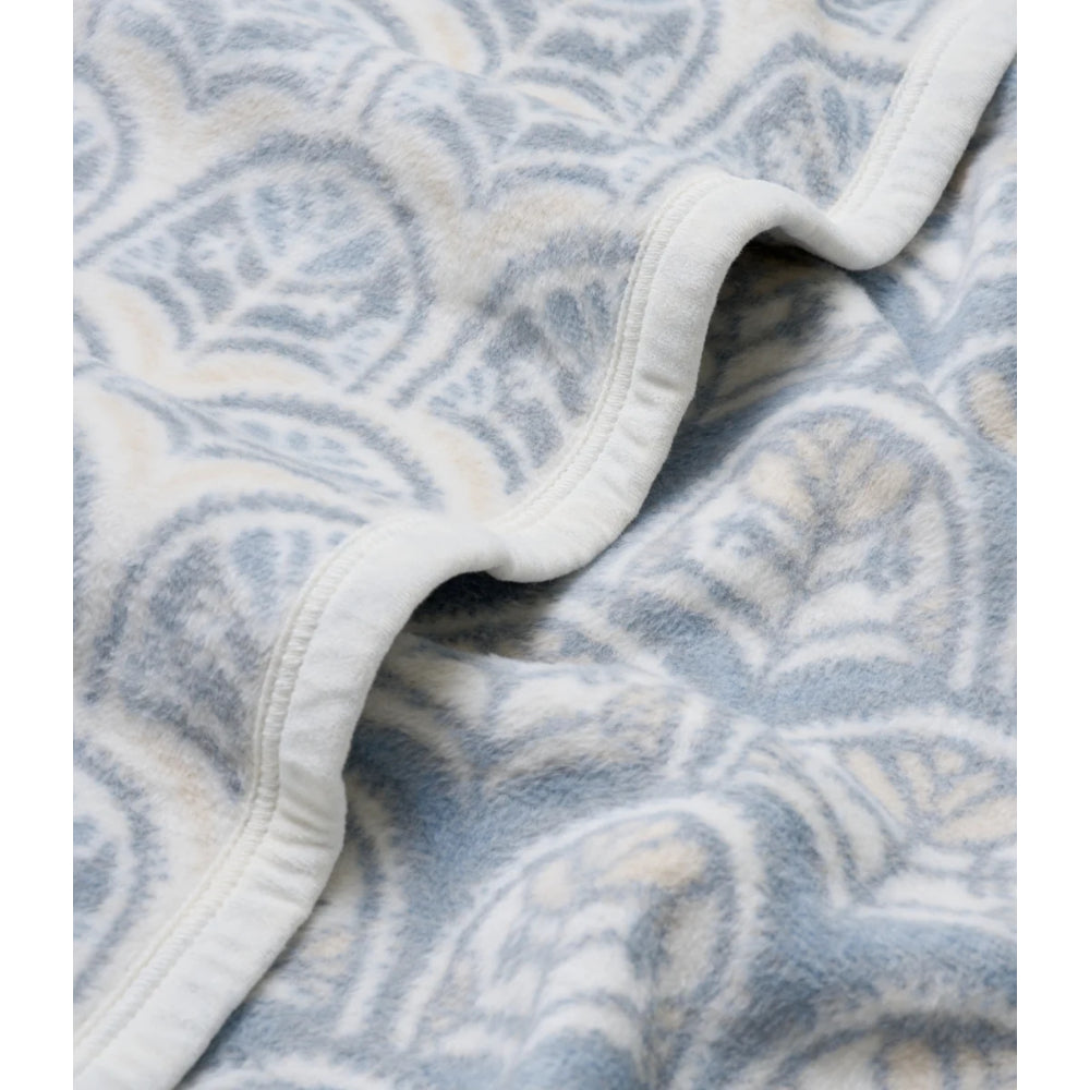 A close-up of a soft fleece blanket with a light blue and cream leafy pattern, folded to show its white stitched edge.