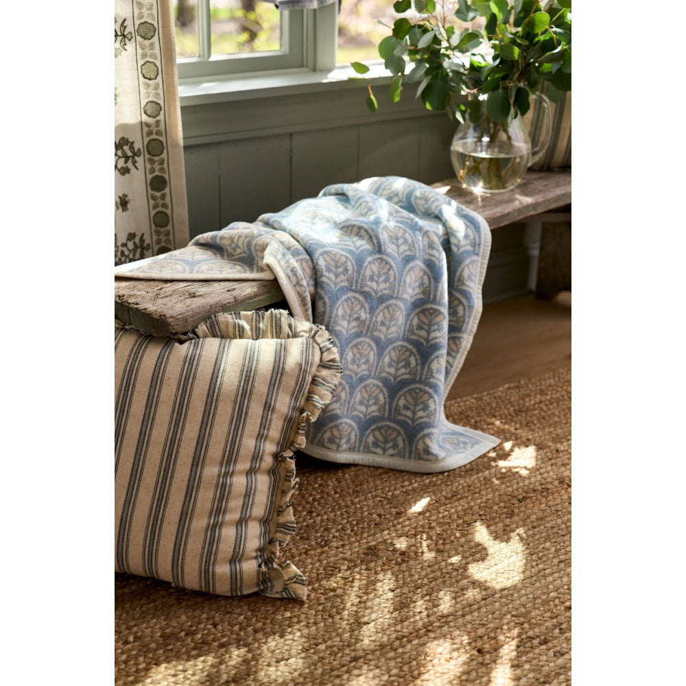 A light blue and beige patterned throw blanket is draped over a rustic wooden bench in a sunlit room, next to a striped beige pillow and a woven rug.