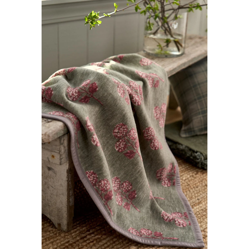 A soft throw blanket draped over a rustic wooden bench, featuring a muted sage green base with a pink floral pattern and a lavender border. The scene is styled with a glass vase of greenery and a plaid cushion in the background.
