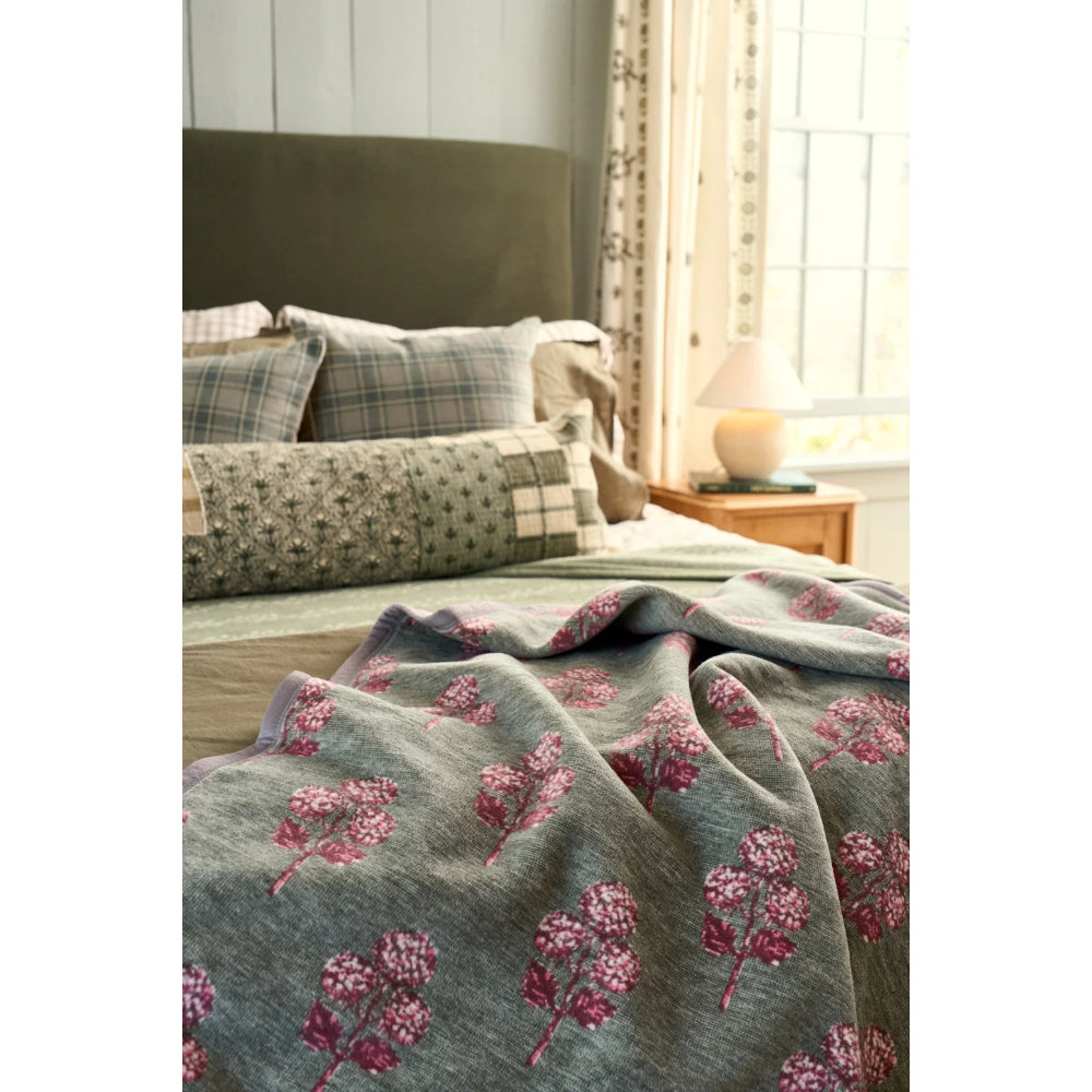 A cozy bedroom scene featuring a textured gray-green throw blanket with a pink floral pattern draped over a bed, styled with plaid and checkered pillows.