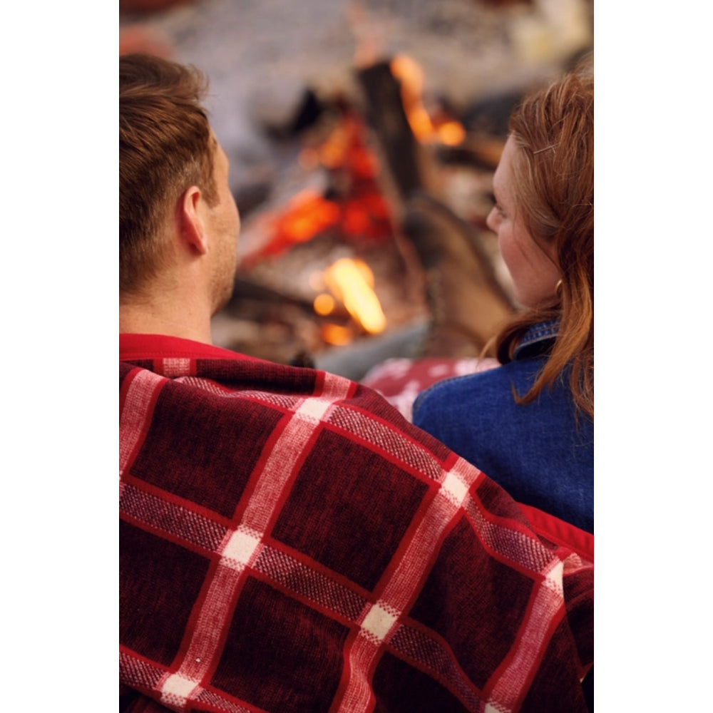 A cozy close-up of a person wrapped in a red and white plaid blanket, sitting next to another person in a blue top in front of a warm fire.