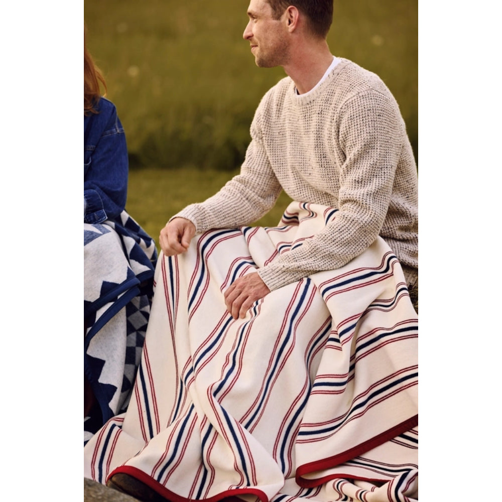A man sitting outdoors wrapped in a large cream blanket with red and navy blue stripes, next to another person holding a blue patterned blanket.
