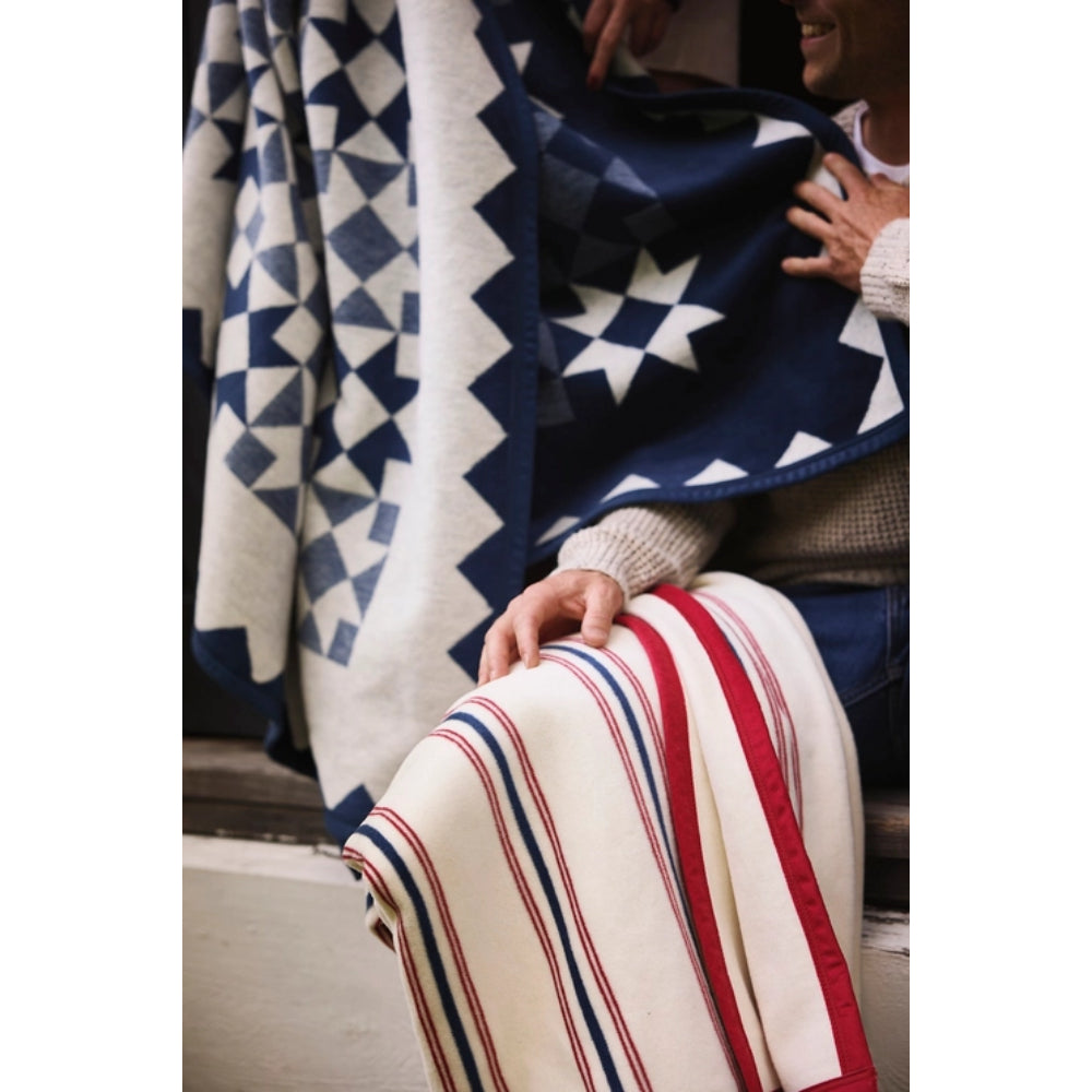 A close-up lifestyle shot of a person wrapped in a patterned blanket. The blanket features a navy blue and cream geometric triangle pattern on one side and a cream base with red and navy blue vertical stripes on the other.
