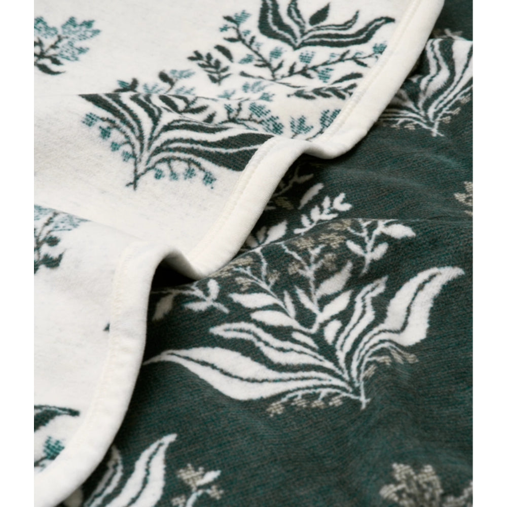 Close-up of a soft blanket with a botanical leaf pattern, showing two overlapping sections: one in off-white with dark green and teal print, and one in dark green with white and gray print.
