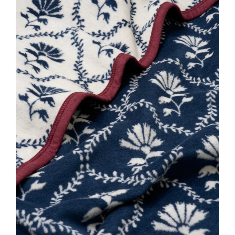 A close-up of a patterned blanket with two visible sides: one side is cream with a dark navy floral and vine print, and the other side is dark navy with a white floral and vine print, separated by a ruffled burgundy edge.
