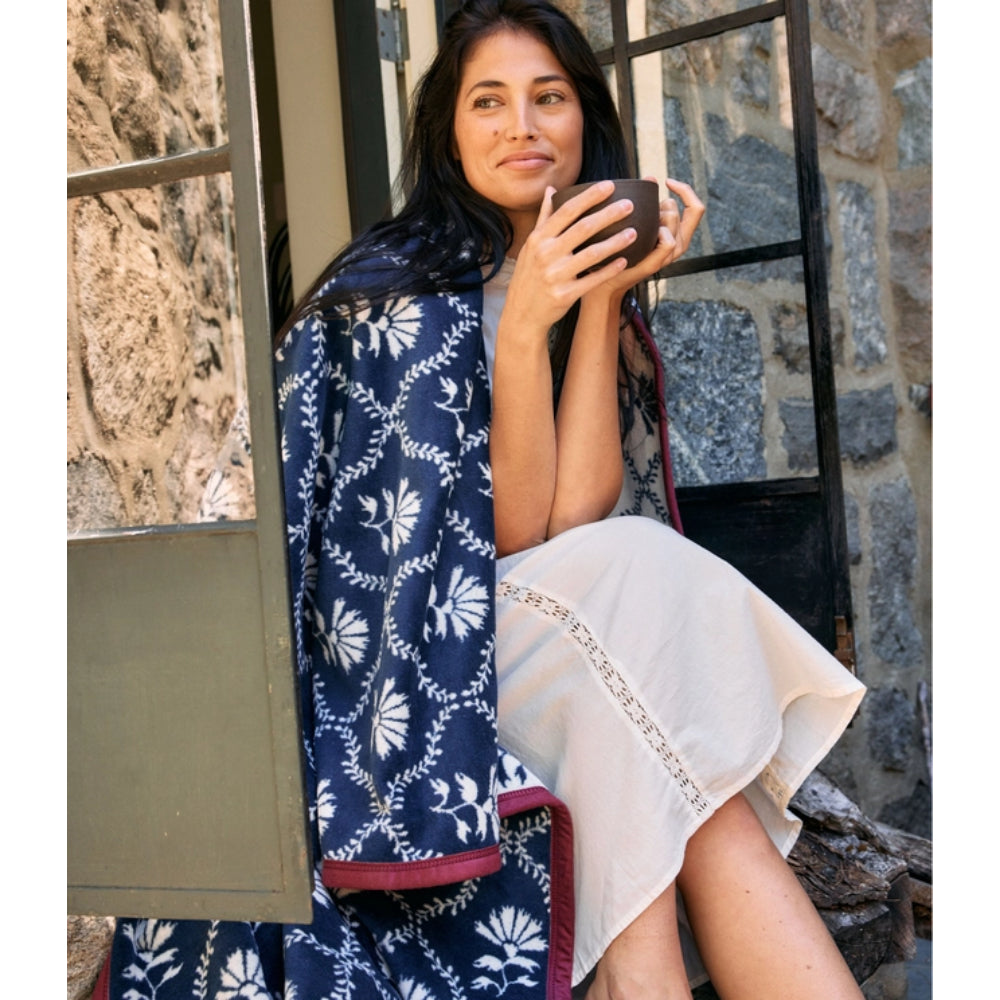 A woman sits outdoors against a stone wall and window, holding a dark cup. She is wrapped around in a navy blue blanket with a white floral and vine pattern and a cream-colored dress.