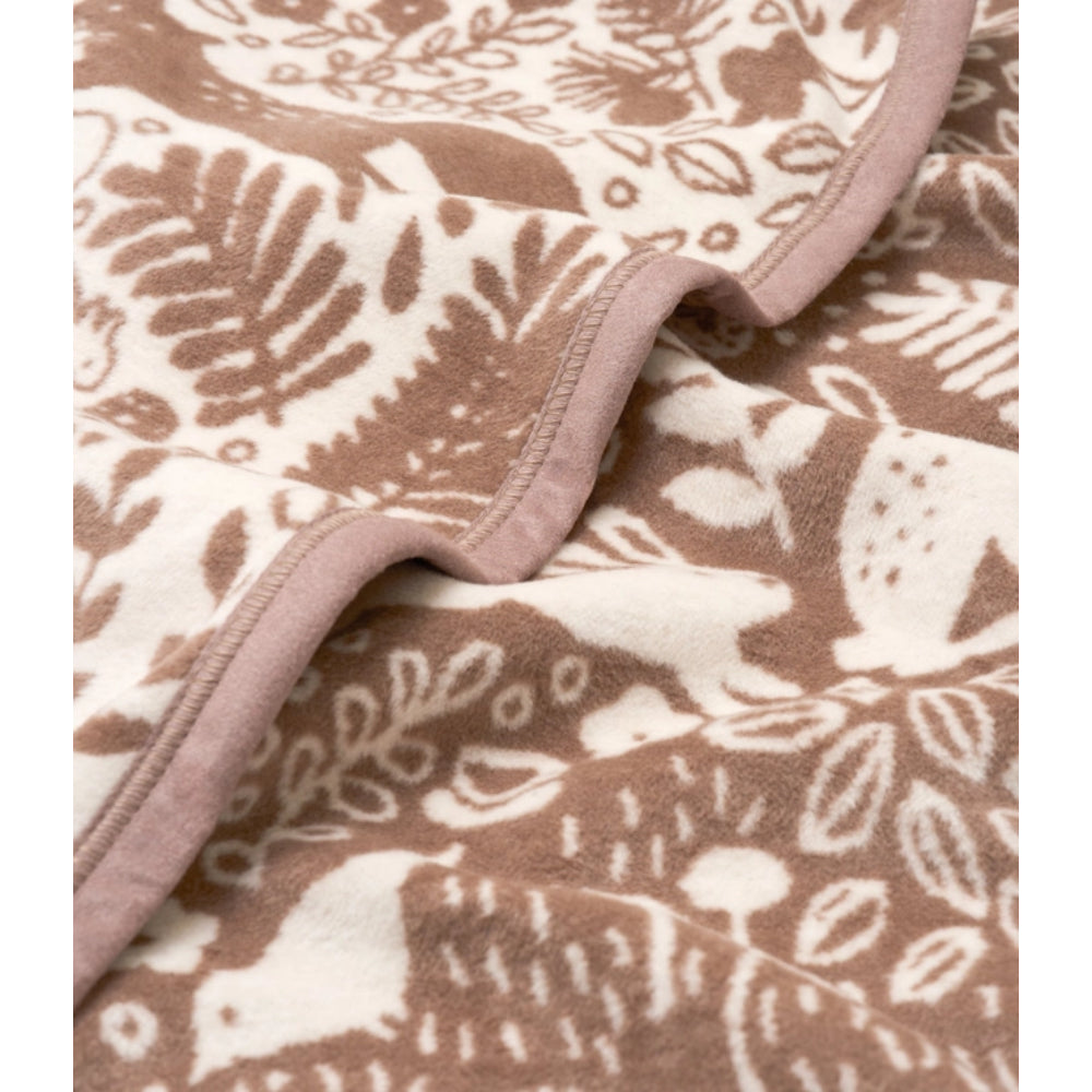 A close-up of a soft fleece blanket with a brown and cream woodland pattern of leaves, birds, and branches, featuring a solid dusty pink ribbed border.