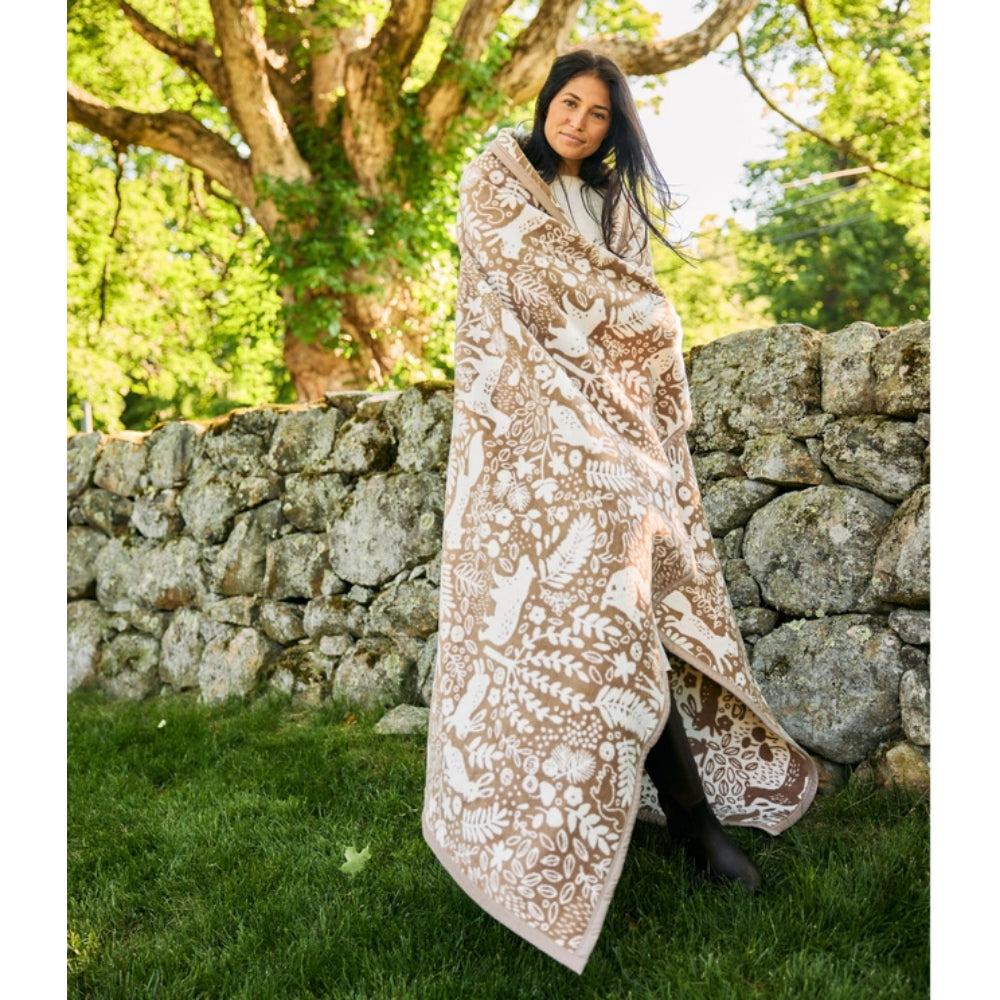 A woman wrapped in a large patterned throw blanket stands outdoors on grass in front of a stone wall and a tree. The blanket has a warm beige base with an intricate white botanical and animal print.