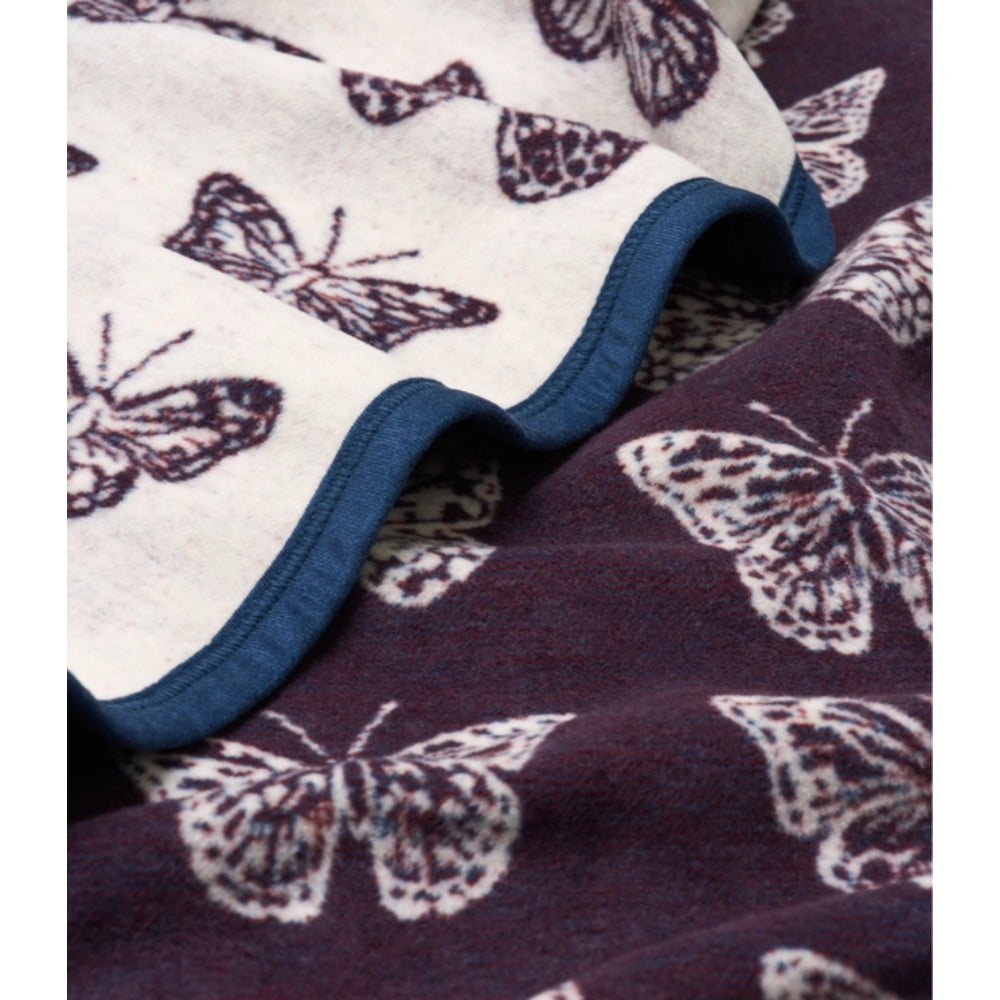 Close-up of a soft blanket showing two sides: one side is off-white with dark butterfly prints and a blue edge, and the other side is dark purple with large white butterfly prints.