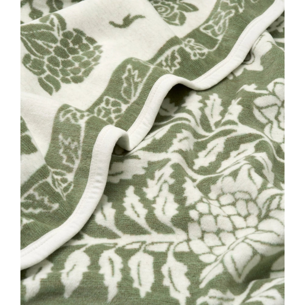 A close-up of a soft blanket with an allover botanical leaf pattern in sage green and off-white, folded to show the fabric texture and white piped edge.