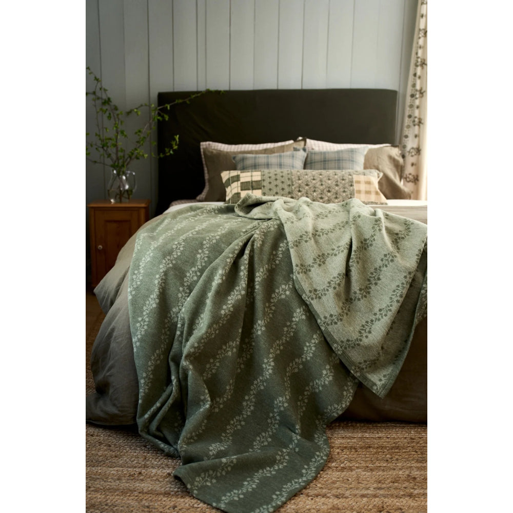 A sage green patterned throw blanket draped over a bed with matching sage green bedding, set in a styled bedroom with a dark headboard, wooden nightstand, and paneled wall.